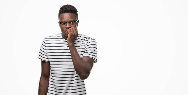 Hombre afroamericano joven con gafas y camiseta azul marino que se ve estresado y nervioso con las manos en la boca mordiendo las uñas. Problema de ansiedad
. - Foto, Imagen