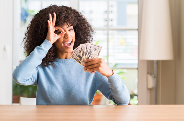 Donna afroamericana in possesso di banconote dollaro con il viso felice sorridente facendo ok segno con mano sugli occhi guardando attraverso le dita
 - Foto, immagini