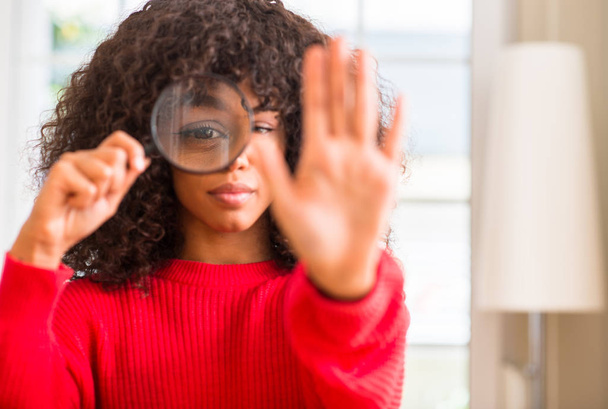 Curiosa mujer afroamericana mirando a través de lupa con la mano abierta haciendo stop sign con expresión seria y segura, gesto de defensa
 - Foto, Imagen
