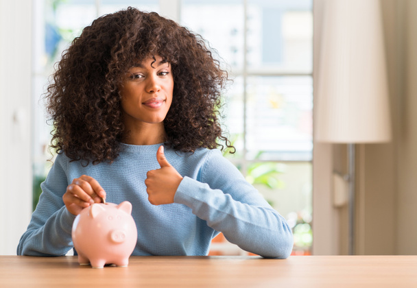 Afrikanische Amerikanerin spart Geld in Sparschwein glücklich mit breitem Lächeln tut ok Zeichen, Daumen nach oben mit Fingern, hervorragende Zeichen - Foto, Bild