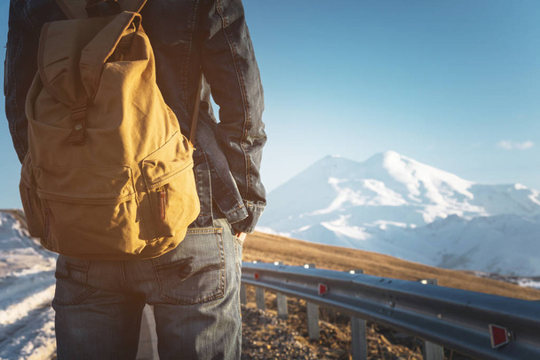 Close-up of a backpack in the back of a male traveler walking along a country road in the background of a mountain. Copy the blank for the dieser. Journey - 写真・画像