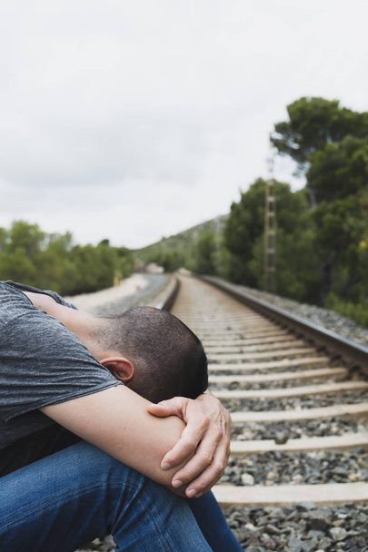 un hombre caucásico, vestido con ropa casual, acurrucado en las vías del ferrocarril, en un paisaje natural
 - Foto, Imagen