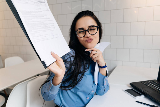 funny young brunette girl with a freelancer in glasses and a blue shirt makes a funny face, holds documents in her hands - Fotó, kép
