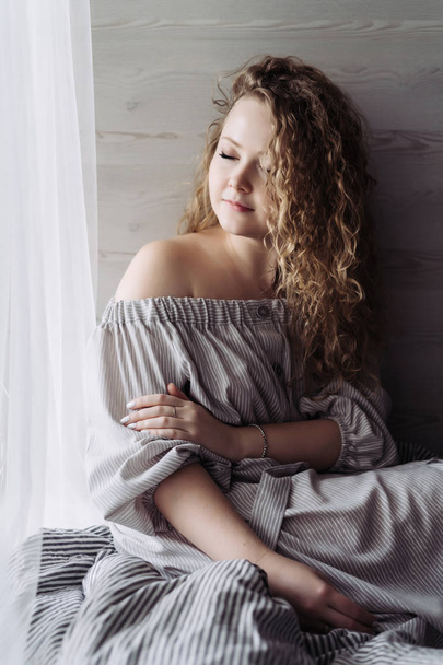 beautiful curly young woman in a cute dress sits by the window, resting, closing her eyes and smiling - Foto, Imagen
