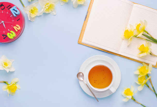 Vazio aberto Notebook Cup of Tea Red Relógio Amarelo Daffodils no fundo azul claro. Mesa de trabalho de mulher com pequeno-almoço acolhedor. Vista superior
 - Foto, Imagem