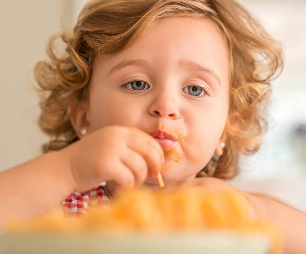 Close up of beautiful blond child eating spaghetti with hands at home. - Фото, зображення