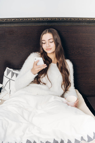 attractive long-haired girl resting in bed late at night, looking into her smartphone - Photo, Image
