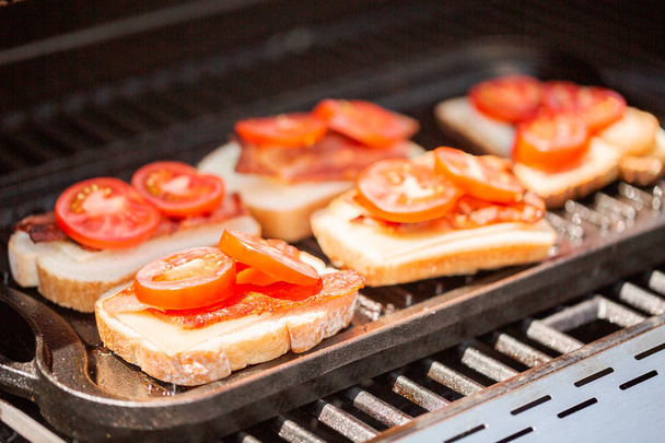 Sándwich de queso a la parrilla con tiras de tocino y tomate fresco en la parrilla de gas al aire libre
. - Foto, Imagen