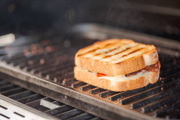 Sándwich de queso a la parrilla con tiras de tocino y tomate fresco en la parrilla de gas al aire libre
. - Foto, Imagen
