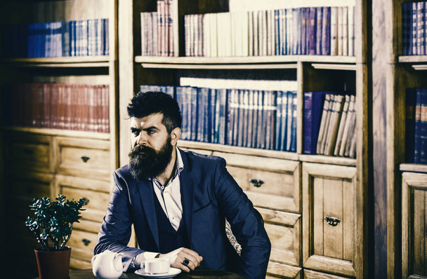 Man in classic suit sits in vintage interior, library, book shelves on background. Oldfashioned man near cup with tea. Aristocratic traditions concept. - Photo, image
