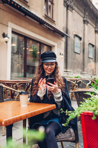 Beautiful young woman having coffee in outdoor cafe while using smartphone. Portrait of stylish smiling girl typing message - Photo, image