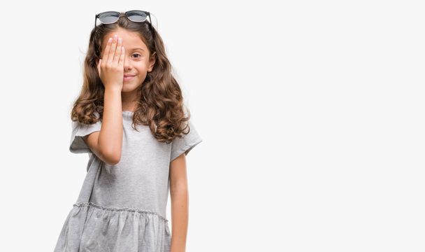 Brunette hispanic girl wearing sunglasses covering one eye with hand with confident smile on face and surprise emotion. - 写真・画像