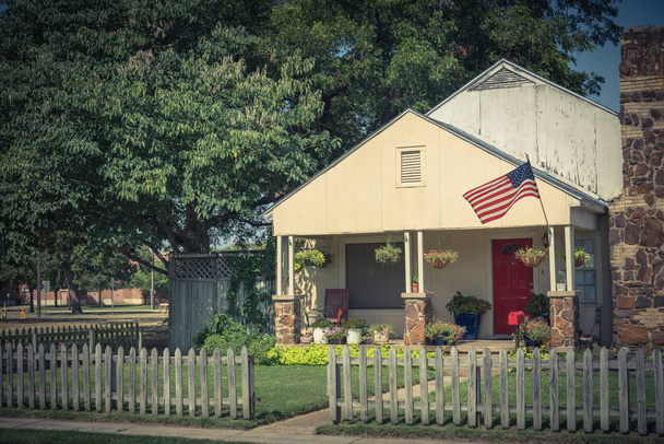 Винтажный скромный дом с шимни в историческом центре города Ирвинг, Техас, США. Классический деревянный забор с ухоженным ландшафтом, цветочные горшки, большое дерево и гордо размахивающий американским флагом
 - Фото, изображение