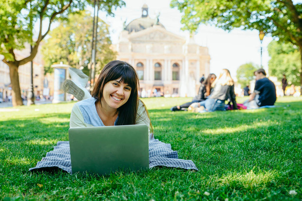 young adult woman laying with laptop in city park on green grass - Foto, afbeelding