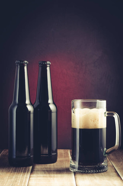 black bottle and glass beer on a dark background with red light/black bottle and glass beer on a dark background with red light. Selective focus and toned - Foto, Imagen