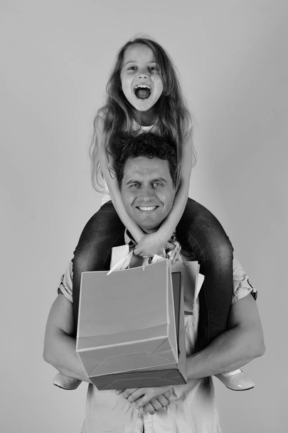 Shopaholics, childhood and family concept. Girl and man with cheerful faces hold shopping bags on pink background. Daughter and father with pink packages. Schoolgirl sits on dads shoulders. - Photo, image