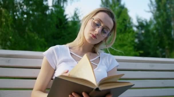 Attractive girl reading a book on the bench and removes hair from her face - Séquence, vidéo