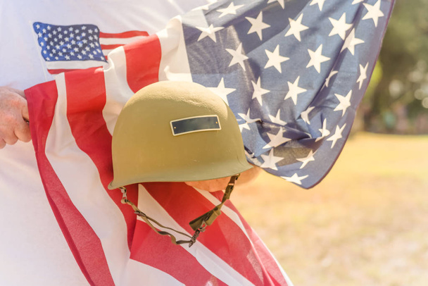 American veteran proudly holding military WWI helmet (M1 helmet) and US flag during parade. July 4th or veteran memorial day poster of WWII, modern wars. Honor and remember soldier troop background - Foto, Imagem