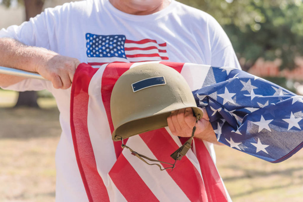 American veteran proudly holding military WWI helmet (M1 helmet) and US flag during parade. July 4th or veteran memorial day poster of WWII, modern wars. Honor and remember soldier troop background - Photo, Image