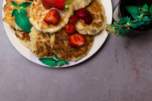 Kwark versgebakken pannenkoeken met rozijnen in een witte plaat, kopje thee met munt, melissa takje op grijze tafel - Foto, afbeelding