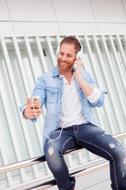 handsome young man wearing denim clothes listening to music via headphones and smartphone in the street - Foto, Bild