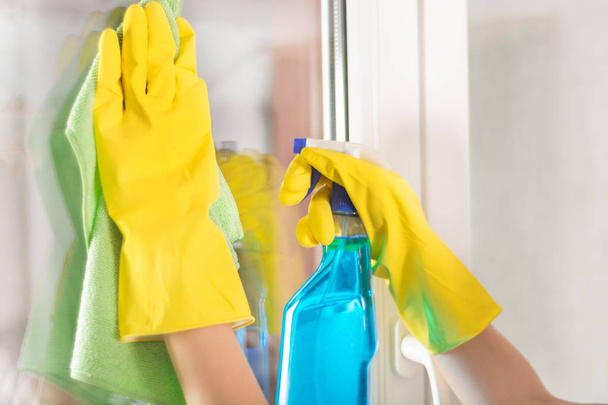 Mains féminines avec des gants de protection jaunes nettoyer la fenêtre à la maison à l'aide de chiffon vert et pulvérisation de détergent. Gros plan, focus sélectif
 - Photo, image