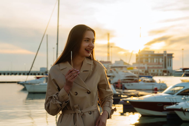 beautiful long-haired smiling girl in trendy beige coat posing in seaport at sunset - Photo, Image