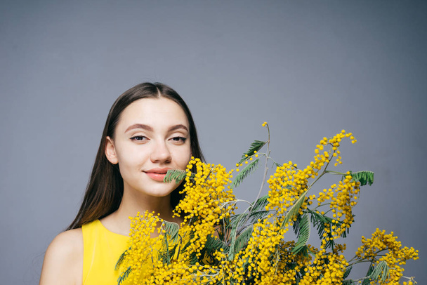 beautiful young girl in yellow dress holds bouquet of fragrant yellow mimosa, posing and smiling - Valokuva, kuva
