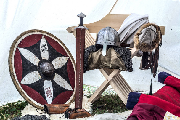 Close up de equipamentos de cavaleiro medieval na velha tenda de dormir. Capacete de metal, escudo, espada e roupas na cadeira. Armas de cavaleiros tradicionais no festival temático da meia-idade
 - Foto, Imagem