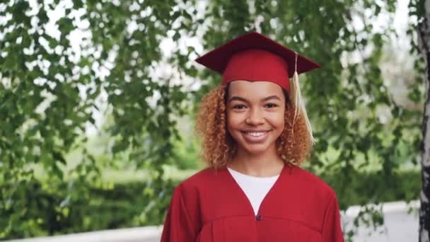 Gülümseyen ve kameraya bakarak oldukça Afro-Amerikan Kız mezun öğrenci kırmızı elbisesi ve kolej açık havada, ayakta portresi. Gençlik ve eğitim kavramı. - Video, Çekim
