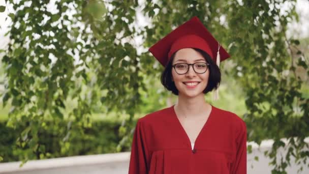 Retrato de jovem alegre estudante de graduação em vestido e placa de argamassa sorrindo e olhando para a câmera de pé sob a árvore no campus. Conceito de juventude e educação
. - Filmagem, Vídeo