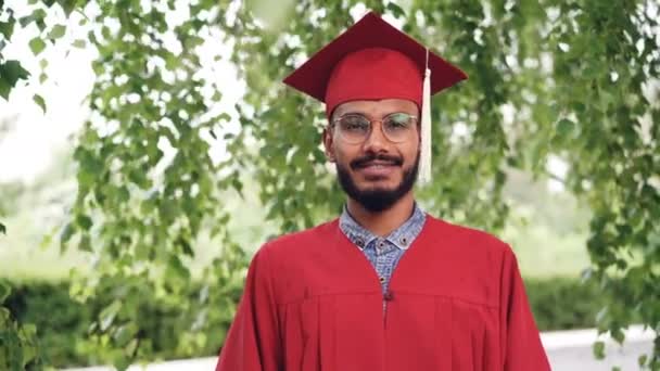 Portrait of bearded mixed race man graduating student in gown and mortar-board smiling and looking at camera standing outdoors on campus. People and education concept. - Footage, Video