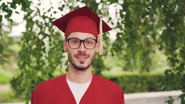 Portrait of bearded young man graduating student wearing glasses, graduation gown and mortar-board smiling and looking at camera standing outdoors. People and education concept. - Footage, Video