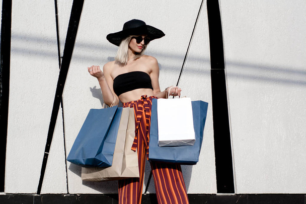 Stylish young woman in sunglasses, hat and pants in a strip is fashionably dressed, goes shopping at an open-air shopping center and poses on a colored background - Photo, image