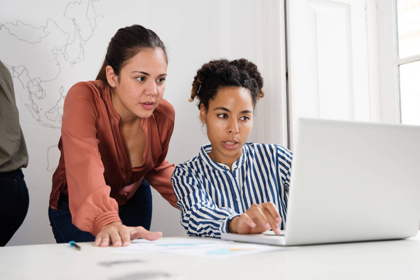 Дві бізнес-леді дивляться на ноутбук на білому столі, в той час як одна жінка друкує на ноутбуці
 - Фото, зображення