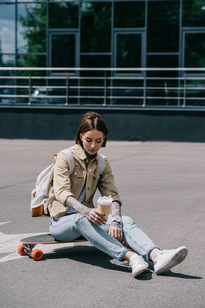 tattooed woman sitting on skateboard with paper cup of coffee  - Zdjęcie, obraz