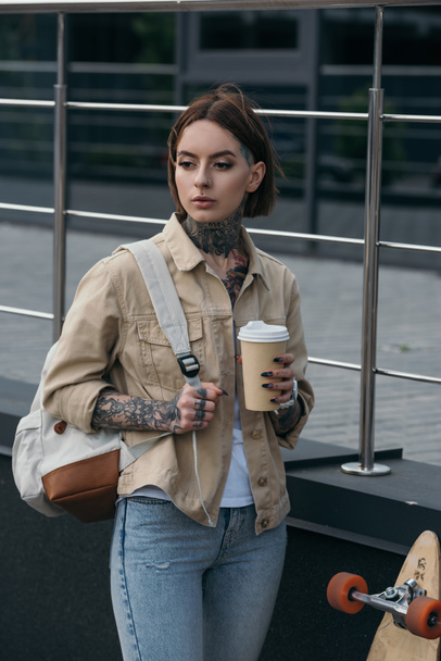 mujer tatuada con mochila sosteniendo taza de café y de pie cerca de monopatín en la calle
  - Foto, Imagen