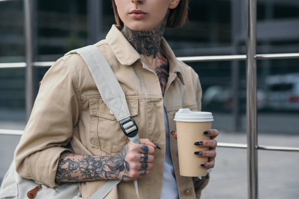 περικομμένη εικόνα κομψή τατουάζ γυναίκα με σακίδιο κρατώντας χαρτί κύπελλο καφέ  - Φωτογραφία, εικόνα