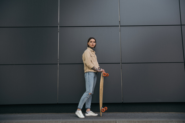 stylish tattooed woman standing with skateboard against black wall  - Fotoğraf, Görsel