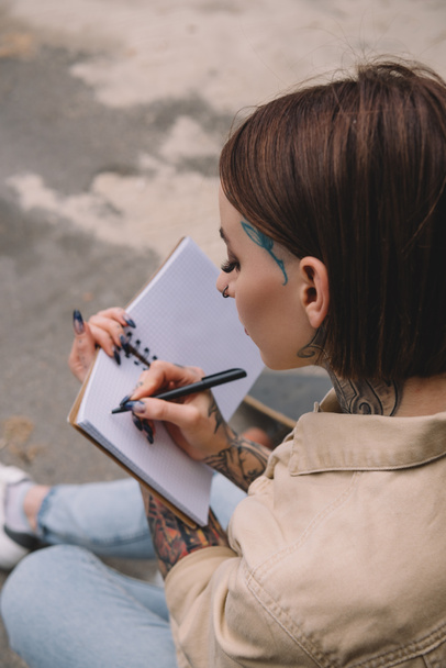 high angle view of tattooed young woman writing in blank notebook  - Photo, Image