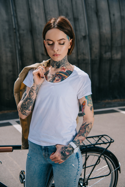 young tattooed woman with closed eyes holding jacket over shoulder near bicycle  - Φωτογραφία, εικόνα