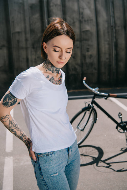 young tattooed woman with closed eyes posing near bicycle at street   - Foto, immagini