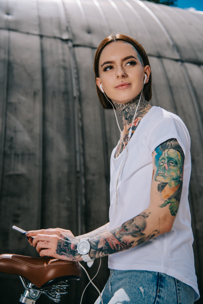 vista de ángulo bajo de mujer tatuada en auriculares escuchando música con teléfono inteligente en bicicleta
 - Foto, Imagen
