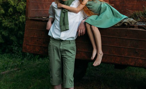 unrequited first love. a barefooted girl in a green, vintage dress, the dress sits on the back of a red, old car and hugs the barefoot neck, next to a standing guy who is dressed in a white shirt, green shorts with a green sweater tied around his nec - Foto, afbeelding