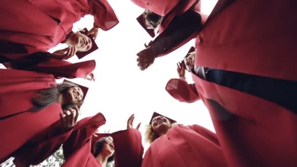 Slow motion low angle shot of graduates standing in circle throwing mortar-boards in the sky and laughing. Togetherness, friendship and modern lifestyle concept. - Footage, Video