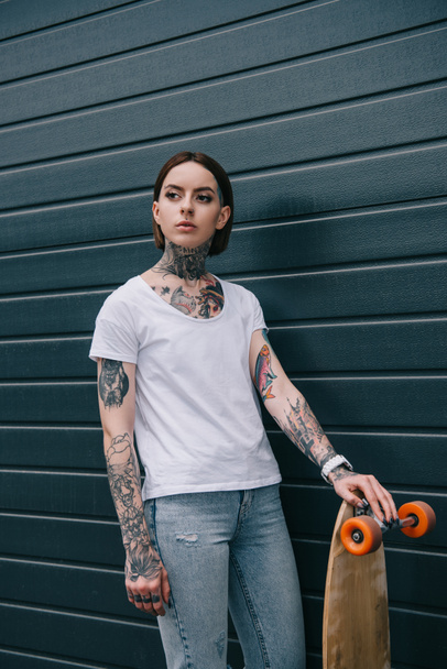 atraktivní vážné Tetovaný dívka stojící s skateboard černé zdi  - Fotografie, Obrázek