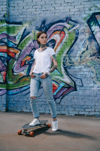 young tattooed woman standing on skateboard near wall with colorful graffiti - 写真・画像