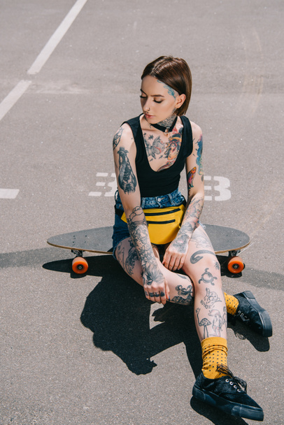 jovem elegante com tatuagens sentado no skate no estacionamento
  - Foto, Imagem