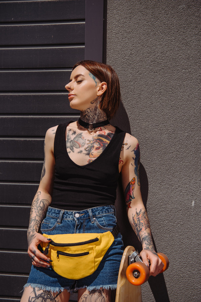 stylish tattooed woman with closed eyes standing with skateboard  - 写真・画像