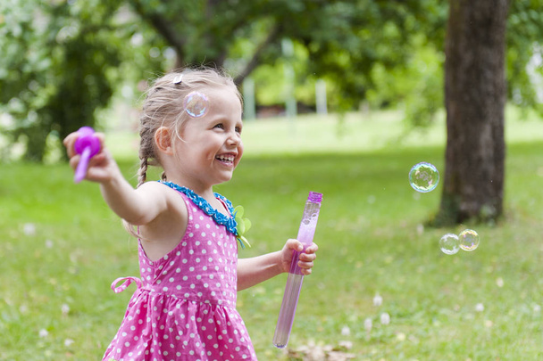 A little cute girl blowing soap bubbles in summer park. - Фото, изображение